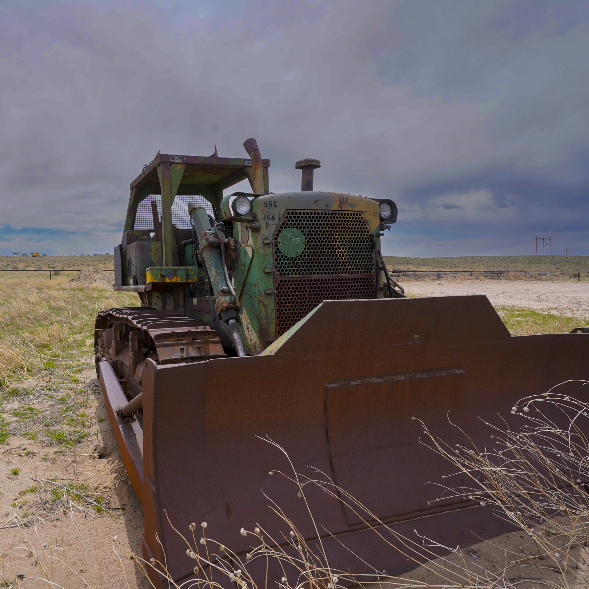 Old, rusted bulldozer with a worn green‑and‑brown exterior, sitting in dry grass with its heavy front blade lowered, under a dark, storm‑clouded sky.